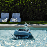 Maytronics Dolphin Eon 100 cordless robotic pool cleaner breaking the water surface in a sunny backyard swimming pool with lounge chairs in the background