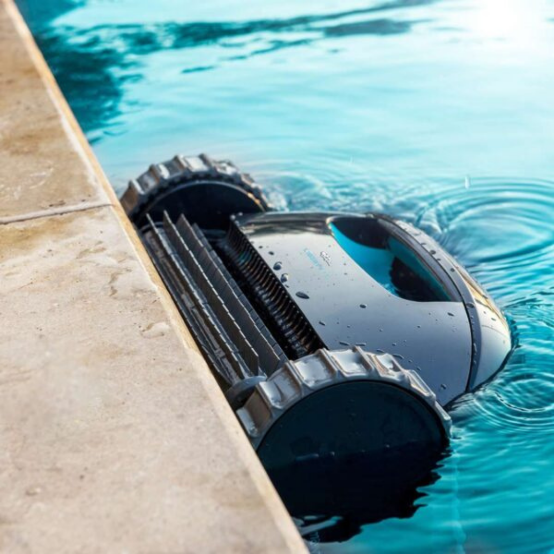 Close-up lifestyle view of the Maytronics Dolphin Liberty 100 robotic pool cleaner scrubbing at the waterline against the pool edge, showing its black body with blue accents, front scrubbing brush, and treaded wheels submerged.