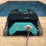 Rear view of the Maytronics Dolphin Liberty 200 robotic pool cleaner resting on a patio next to a swimming pool, showing its teal rear panel with power button and magnetic induction charger cable attached.