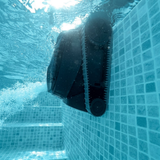 Underwater action shot looking up at the Maytronics Dolphin Liberty 200 robotic pool cleaner as it climbs vertical mosaic pool tiles while expelling a stream of water from its rear propulsion jet near the surface.
