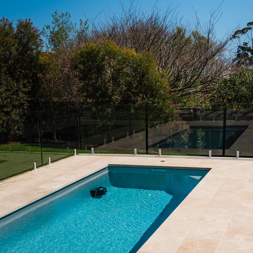 High-angle lifestyle shot of an outdoor backyard swimming pool with glass fencing, showing the Maytronics Dolphin Liberty 400 robotic pool cleaner operating along the wall underwater.