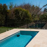 High-angle lifestyle shot of an outdoor backyard swimming pool with glass fencing, showing the Maytronics Dolphin Liberty 400 robotic pool cleaner operating along the wall underwater.