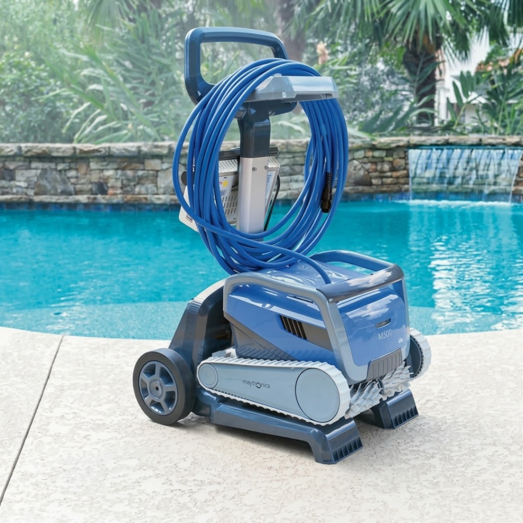 Three-quarter lifestyle view of a blue Maytronics Dolphin M500 robotic pool cleaner resting on its transport caddy on a paved pool deck, featuring its light grey tracks, white scrubbing brushes, coiled blue floating cable, power supply unit, and a swimming pool with stone waterfall backdrop.
