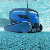 Underwater side-angle action shot of the blue Maytronics Dolphin M 600 robotic pool cleaner driving along a pebblecrete pool floor, displaying its large side tracks and active front scrubbing brushes.