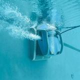 Underwater side profile view of the blue Maytronics Dolphin M 600 robotic pool cleaner scrubbing along a vertical pool wall near the surface, expelling a plume of air bubbles and water jets.
