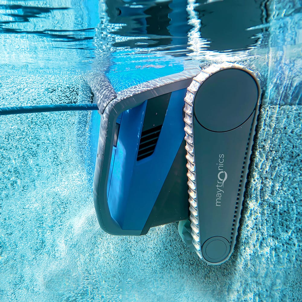 Close-up underwater action shot of the blue Maytronics Dolphin M 600 robotic pool cleaner scrubbing sideways along the pool tile line at the water surface, creating air bubbles along its tracks.