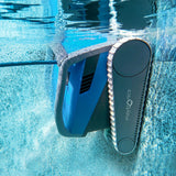 Close-up underwater action shot of the blue Maytronics Dolphin M 600 robotic pool cleaner scrubbing sideways along the pool tile line at the water surface, creating air bubbles along its tracks.