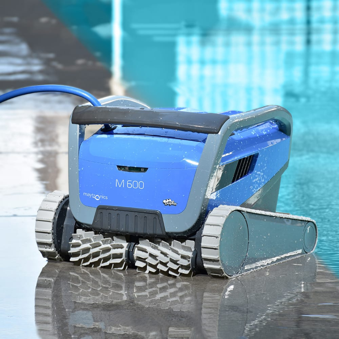 Low-angle lifestyle view of the wet Maytronics Dolphin M 600 robotic pool cleaner sitting poolside with water reflections, showcasing its blue body, front active scrubbing brushes, grey tracks, and blue power cable.