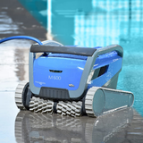 Low-angle lifestyle view of the wet Maytronics Dolphin M 600 robotic pool cleaner sitting poolside with water reflections, showcasing its blue body, front active scrubbing brushes, grey tracks, and blue power cable.