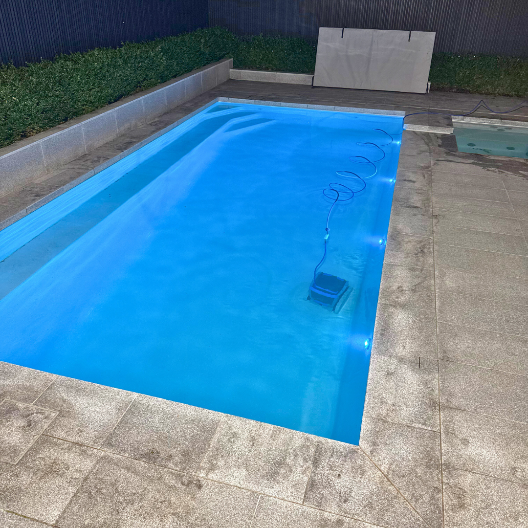 Wide-angle outdoor lifestyle view of an in-ground swimming pool surrounded by stone pavers, with a Maytronics Dolphin M 700 robotic pool cleaner actively operating at the bottom of the clear blue water.