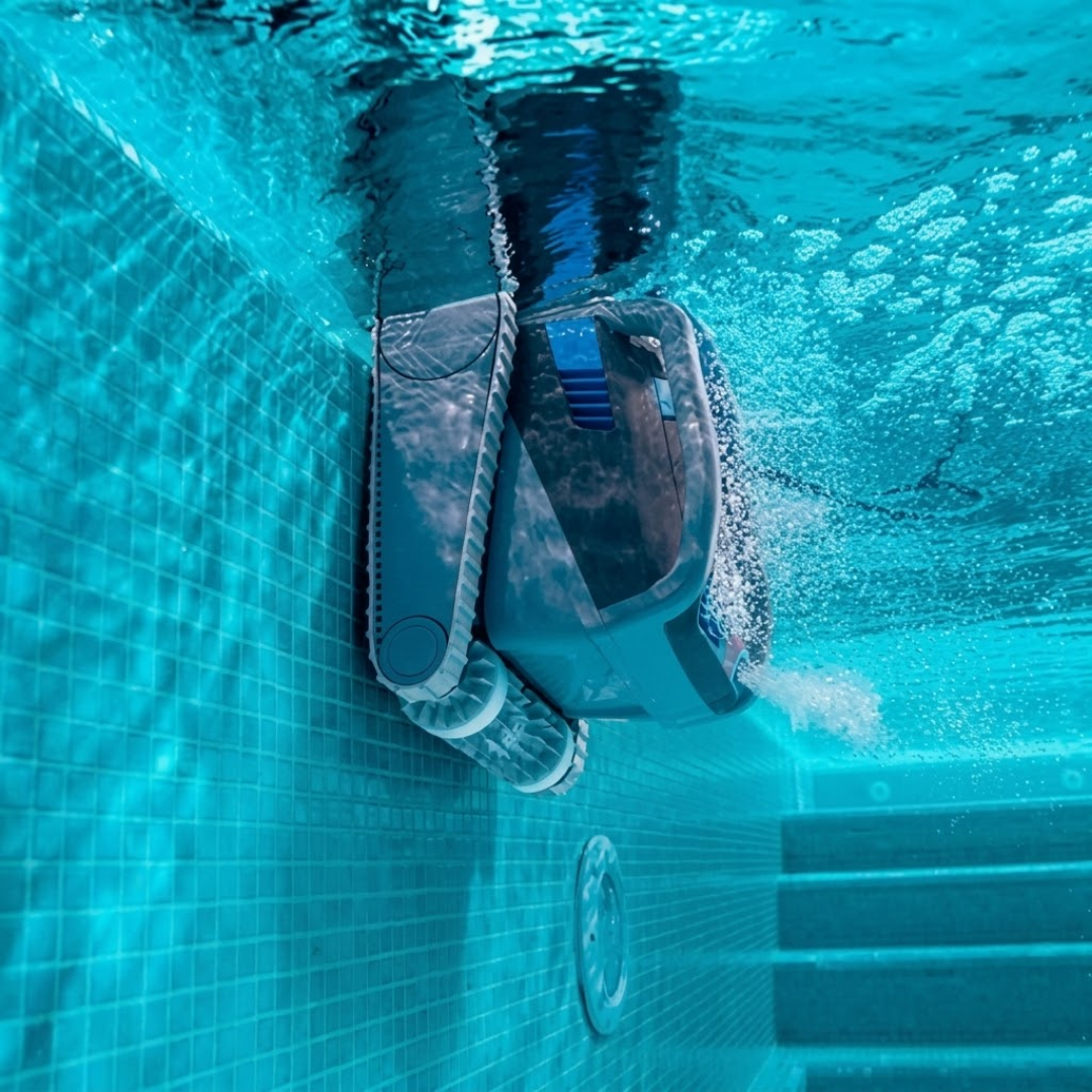 Underwater profile view of the dark grey Maytronics Dolphin M 700 robotic pool cleaner climbing a blue tiled pool wall near the surface, releasing a steady stream of air bubbles.