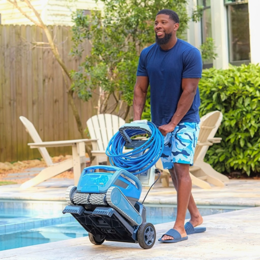 Outdoor lifestyle photo of a smiling man wheeling a light blue Maytronics Dolphin robotic pool cleaner on its transport caddy along a paved poolside patio, displaying the coiled blue floating cable, power supply unit, and outdoor patio furniture in the background.