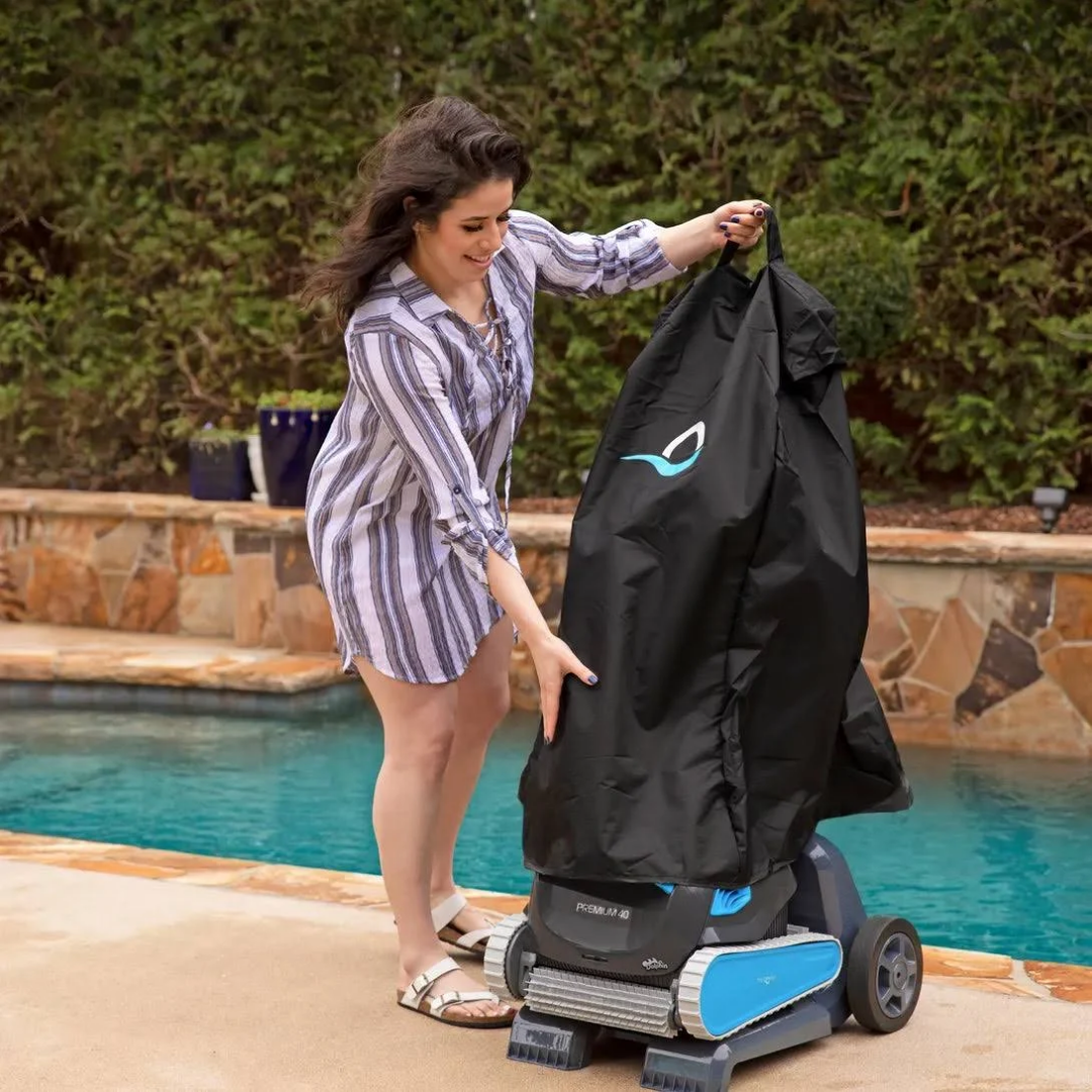 Outdoor lifestyle photo of a woman fitting a black weather-resistant protective cover onto a Maytronics Dolphin pool cleaner mounted on its transport caddy, located on a stone pool deck alongside a swimming pool.