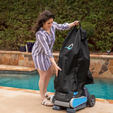 Outdoor lifestyle photo of a woman fitting a black weather-resistant protective cover onto a Maytronics Dolphin pool cleaner mounted on its transport caddy, located on a stone pool deck alongside a swimming pool.