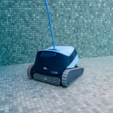 Underwater front three-quarter view of the Maytronics Dolphin S1000 robotic pool cleaner resting on a blue mosaic tiled pool floor near the pool wall, featuring its front active brush, rubber track drive system, S1000 model branding, and blue power cable rising to the surface.