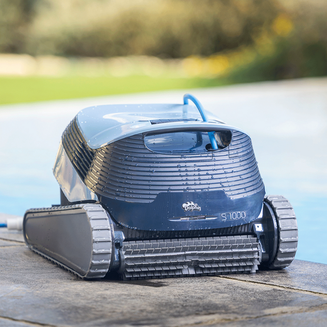 Low-angle outdoor lifestyle view of the Maytronics Dolphin S1000 robotic pool cleaner resting wet on stone coping by the pool edge, showing its front scrubbing brush, rubber continuous tracks, dark grey body with water droplets, slate blue top hatch, and Dolphin S1000 branding.