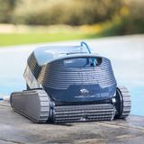 Low-angle outdoor lifestyle view of the Maytronics Dolphin S1000 robotic pool cleaner resting wet on stone coping by the pool edge, showing its front scrubbing brush, rubber continuous tracks, dark grey body with water droplets, slate blue top hatch, and Dolphin S1000 branding.