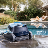 Outdoor lifestyle view of the Maytronics Dolphin S1000 robotic pool cleaner resting wet on a dark stone pool deck, with its power cable extending into the water and a person relaxing on a sun lounger in the blurred background.
