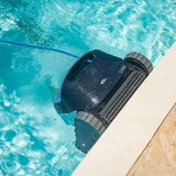 High-angle outdoor lifestyle view of the Maytronics Dolphin S1000 robotic pool cleaner climbing and scrubbing the pool wall at the waterline next to stone pool coping, showcasing its active brush, track wheels, blue power cable, and S1000 model branding.