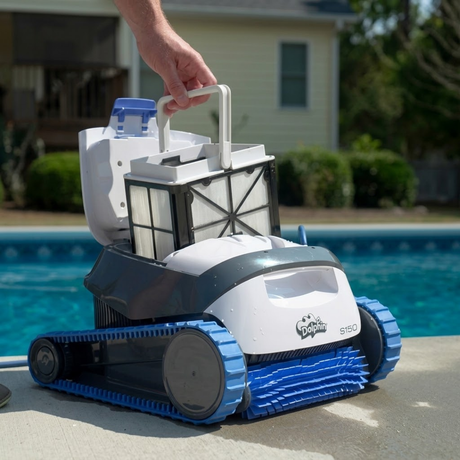 Outdoor lifestyle view of a hand lifting the top-access fine filter basket out of the white and dark grey Maytronics Dolphin S 150 robotic pool cleaner resting on concrete poolside coping.