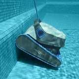 Underwater side view of the white and dark grey Maytronics Dolphin S 150 robotic pool cleaner navigating and cleaning pool steps lined with blue tiles.