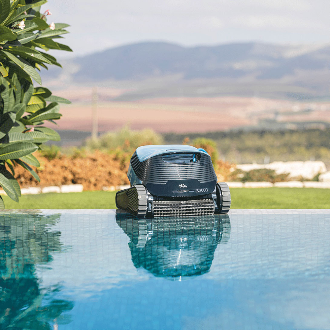 Wide-angle outdoor lifestyle view of the Maytronics Dolphin S2000 robotic pool cleaner resting at the edge of an infinity pool with its reflection visible in the calm water, set against a scenic backdrop of rolling hills and lush vegetation.