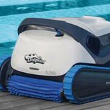 Close-up outdoor lifestyle view of the white and dark grey Maytronics Dolphin S 250 robotic pool cleaner resting on a wooden deck by the pool, detailing its blue active front scrubbing brush and continuous tracks.