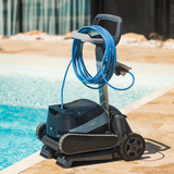 Outdoor lifestyle view of the Maytronics Dolphin S3000 robotic pool cleaner mounted on its premium transport caddy by the poolside, showing the coiled blue power cable neatly wrapped on the caddy hook and power supply unit.