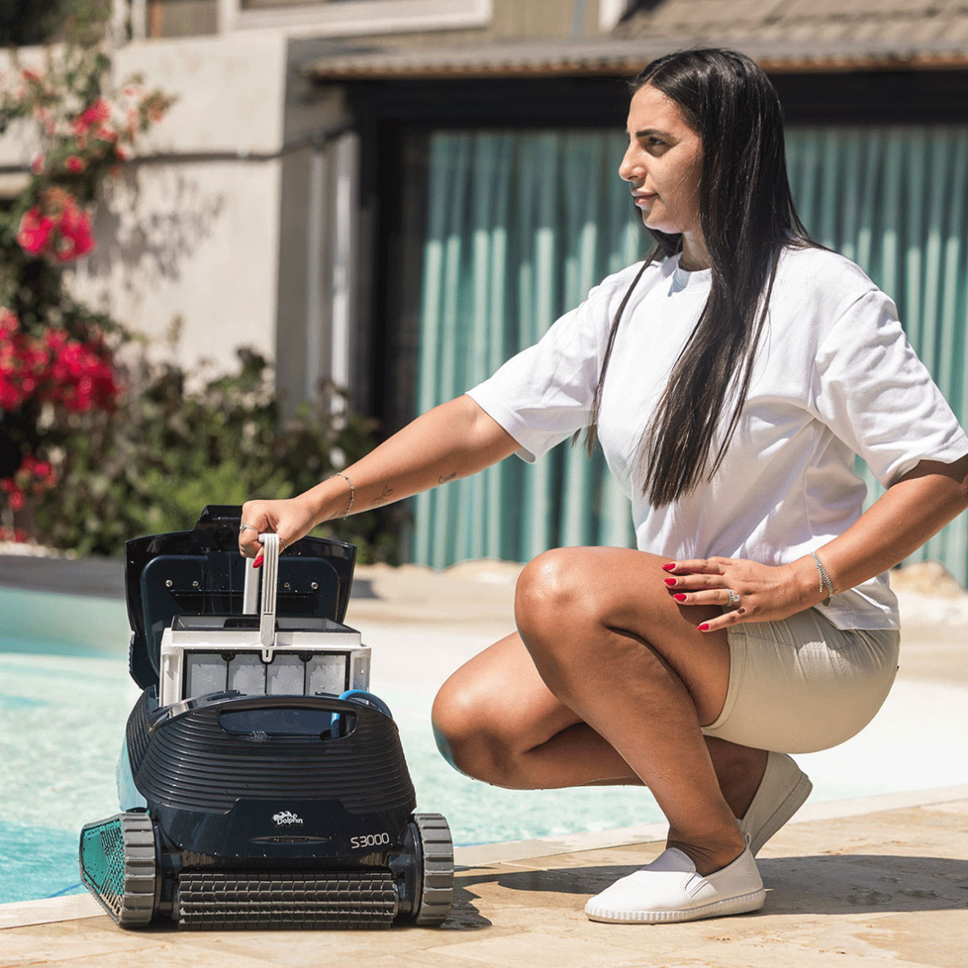 Outdoor lifestyle view of a woman crouching by the pool lifting the large top-access filter basket out of the open Maytronics Dolphin S3000 robotic pool cleaner, demonstrating easy maintenance and cleaning.