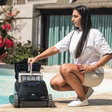 Outdoor lifestyle view of a woman crouching by the pool lifting the large top-access filter basket out of the open Maytronics Dolphin S3000 robotic pool cleaner, demonstrating easy maintenance and cleaning.