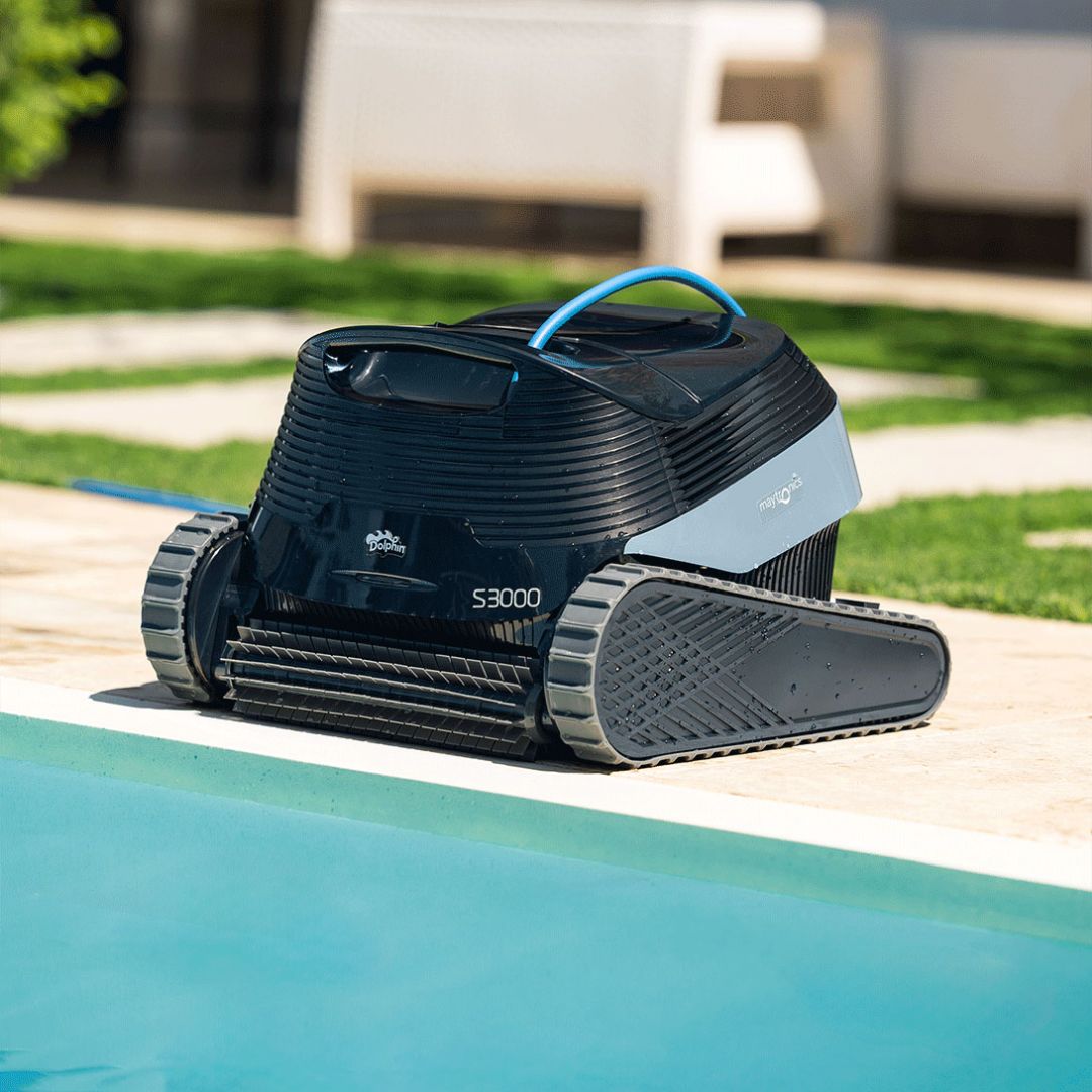 Low-angle outdoor lifestyle view of the Maytronics Dolphin S3000 robotic pool cleaner resting on stone poolside paving by clear blue water, showcasing its front active scrubbing brush, rubber continuous tracks, dark navy chassis, slate blue side panel with Maytronics branding, Dolphin S3000 logo, and blue top handle.