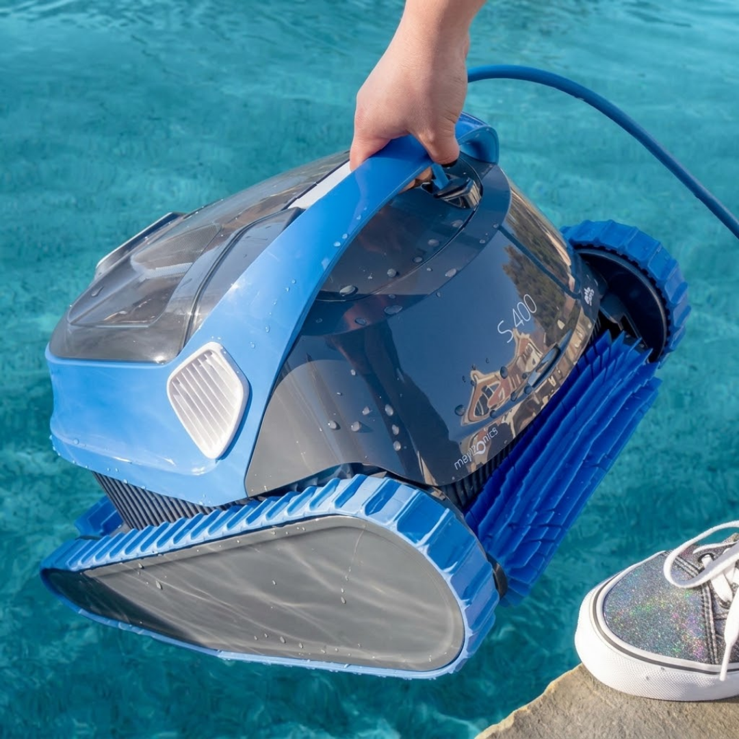 Close-up poolside lifestyle view of a hand lifting the dark grey and blue Maytronics Dolphin S 400 robotic pool cleaner out of the water by its blue ergonomic handle.