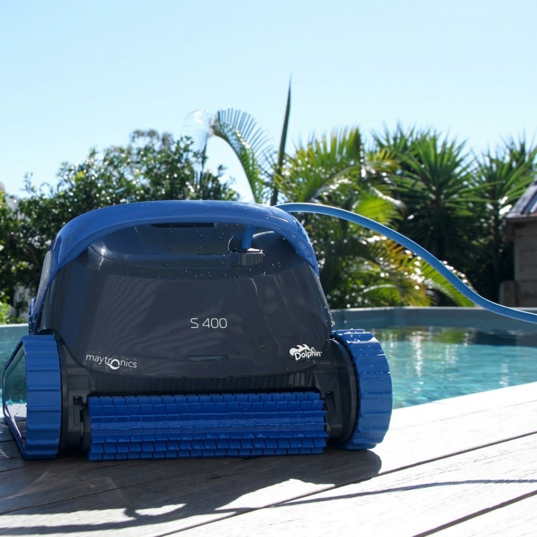 Outdoor lifestyle front view of the dark grey and blue Maytronics Dolphin S 400 robotic pool cleaner resting on a wooden pool deck next to a sunny swimming pool.