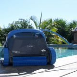 Outdoor lifestyle front view of the dark grey and blue Maytronics Dolphin S 400 robotic pool cleaner resting on a wooden pool deck next to a sunny swimming pool.