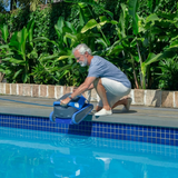 Outdoor lifestyle view of a man kneeling poolside to lower the dark grey and blue Maytronics Dolphin S 400 robotic pool cleaner into a clear blue swimming pool.