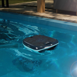 Lifestyle view of the Maytronics Dolphin Skimmi 200 robotic pool skimmer floating and operating in clear blue pool water alongside a patio deck, showcasing its top circuit-patterned solar panel and dark grey shell with teal trim.