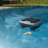 Lifestyle view of the Maytronics Dolphin Skimmi 300 robotic pool skimmer floating on clear blue pool water approaching floating leaves, showcasing its slate blue upper body, dark lower chassis, top solar panel, and front Dolphin logo.