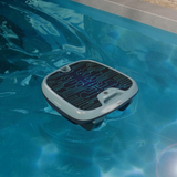 High-angle lifestyle view of the Maytronics Dolphin Skimmi 300 robotic pool skimmer floating on sunlight-dappled pool water near the coping, highlighting its top circuit-board pattern solar panel, slate blue trim, and front control module.
