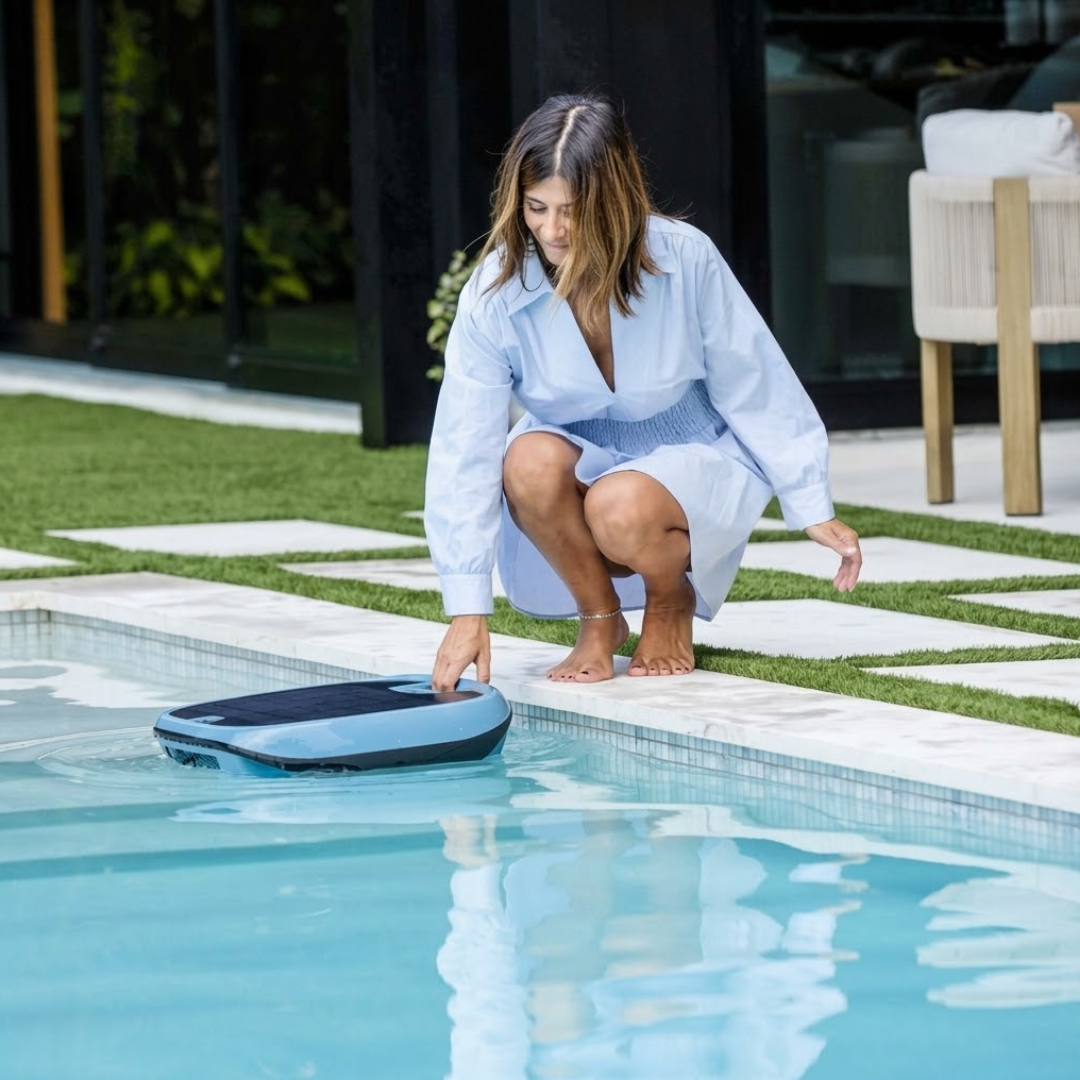 Lifestyle image of a woman crouching poolside, placing the Maytronics Dolphin Skimmi 300 robotic pool skimmer into the water by its integrated top handle next to a stone deck and lawn area.