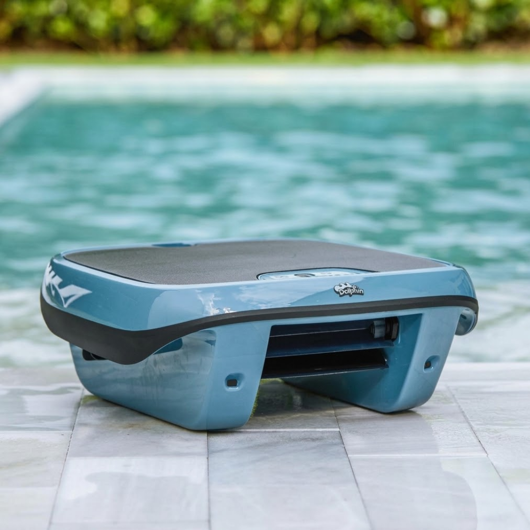 Outdoor lifestyle shot of the Maytronics Dolphin Skimmi 300 robotic pool skimmer resting poolside on white stone patio tile, showing its slate blue chassis, dark trim band, front debris intake mouth, and dark solar panel top with the pool in the background.