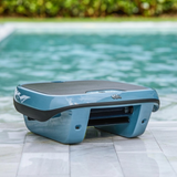 Outdoor lifestyle shot of the Maytronics Dolphin Skimmi 300 robotic pool skimmer resting poolside on white stone patio tile, showing its slate blue chassis, dark trim band, front debris intake mouth, and dark solar panel top with the pool in the background.