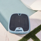 Top-down outdoor lifestyle view of the Maytronics Dolphin Skimmi 300 robotic pool skimmer resting at the pool edge on stone coping near a grass inlay, highlighting its grid-patterned solar panel top, slate blue housing, handle release latch, and front control module.
