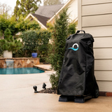 Outdoor lifestyle photo of a Maytronics Dolphin pool cleaner transport caddy protected by its black weather-resistant cover, standing on a paved patio beside a residential house wall with a swimming pool and landscaped garden in the background.