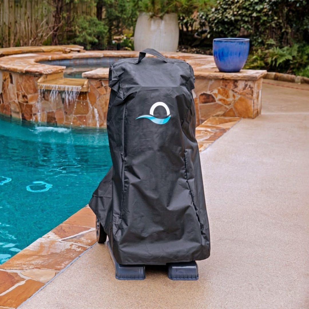 Outdoor lifestyle view of a black weather-resistant protective cover fitted over a Maytronics Dolphin pool cleaner transport caddy on a paved pool deck, featuring the blue Maytronics wave logo, top carrying handle, and a swimming pool with a stone waterfall in the background.