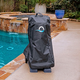 Outdoor lifestyle view of a black weather-resistant protective cover fitted over a Maytronics Dolphin pool cleaner transport caddy on a paved pool deck, featuring the blue Maytronics wave logo, top carrying handle, and a swimming pool with a stone waterfall in the background.