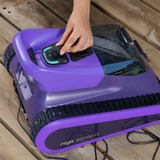 Close-up lifestyle view of a hand connecting the charging cable to the top-mounted charging port of the purple-accented Maytronics Niya Sonar F3 robotic pool cleaner resting on a wooden deck.