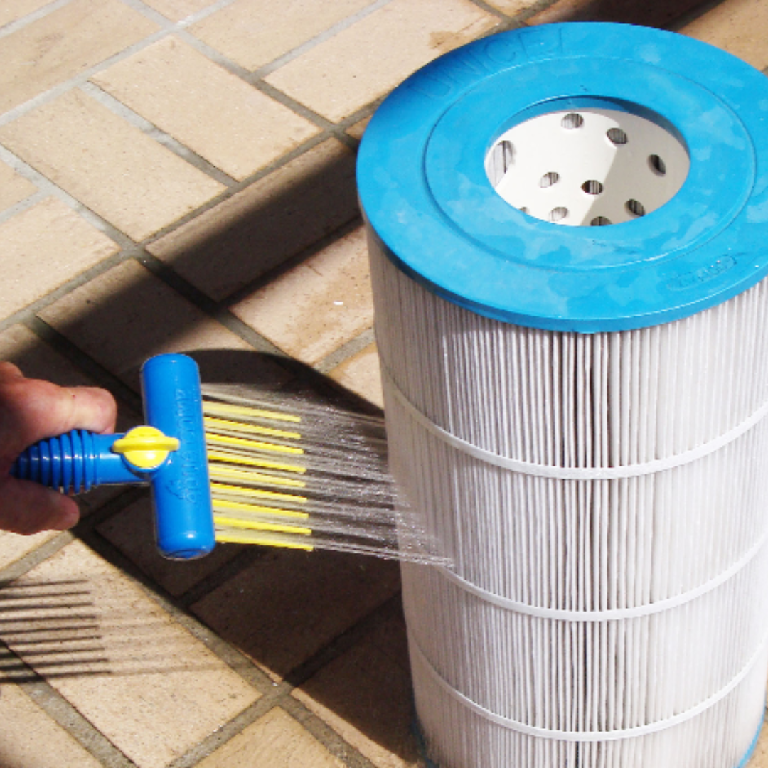 A hand holding a blue and yellow Pool Aqua Comb tool, spraying high-pressure water streams directly into the pleats of a large white and blue Unicel swimming pool cartridge filter on a tiled patio.