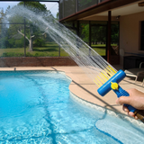 Action lifestyle photo of a hand holding a blue Pool AquaComb tool spraying a wide, powerful fan of water from its yellow comb fingers across a backyard swimming pool setting.