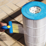 Action photo of a hand holding a blue Pool AquaComb tool spraying targeted water jets through yellow comb teeth into a white Unicel pool cartridge filter, set against a outdoor tiled ground background.