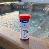 A bottle of Poppits Peroxsil Test Strips sitting on the acrylic edge of an outdoor spa, used for monitoring hydrogen peroxide, alkalinity, and pH levels in chlorine-free hot tubs.